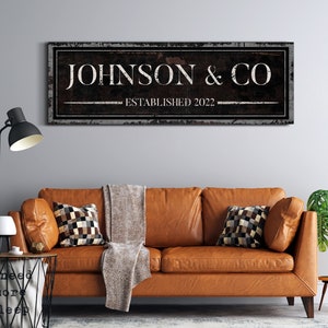 May include: A black and white sign with a distressed look, featuring the text "JOHNSON & CO ESTABLISHED 2022". The sign is hanging on a gray wall above a brown leather couch.