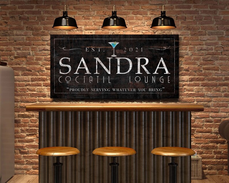 May include: A custom-made sign for a cocktail lounge, featuring the name "SANDRA" in large white letters. The sign includes the text "COCKTAIL LOUNGE" and "Proudly serving whatever you bring" on a dark background, with a martini glass graphic.