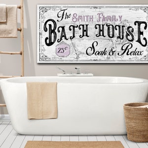May include: White bathroom sign with black text that reads "The Smith Family Bath House 25&cent; Soak & Relax". The sign is hanging above a white bathtub with a beige towel hanging on the side.