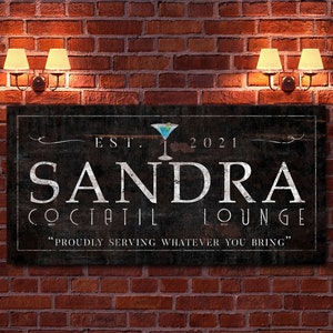 May include: A rectangular wooden sign with the words "SANDRA COCKTAIL LOUNGE" in white lettering. The sign includes a martini glass graphic and the text "EST. 2021". The sign is mounted on a red brick wall.