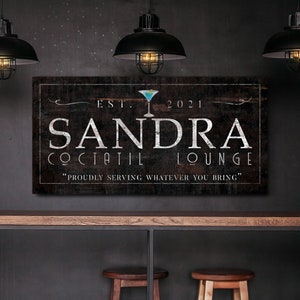 May include: A rectangular wooden sign with the text "SANDRA COCTAIL LOUNGE" in white lettering. The sign includes the year "2021" and a cocktail glass graphic. The phrase "PROUDLY SERVING WHATEVER YOU BRING" is below the main text.