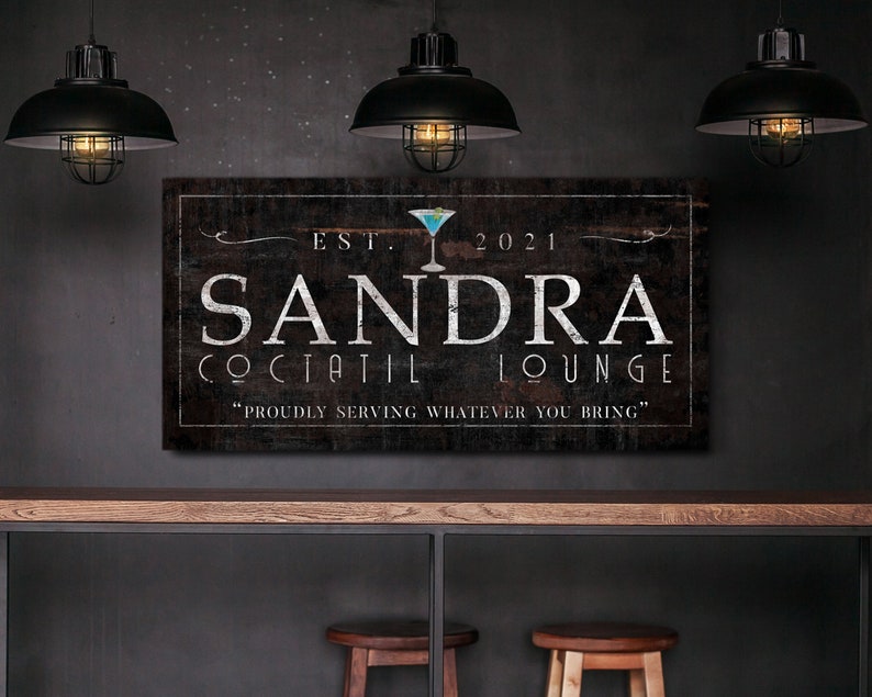 May include: A rectangular wooden sign with the text "SANDRA COCTAIL LOUNGE" in white lettering. The sign includes the year "2021" and a cocktail glass graphic. The phrase "PROUDLY SERVING WHATEVER YOU BRING" is below the main text.
