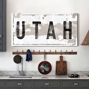 May include: A white canvas wall art print with black text that reads "UTAH" in a distressed font. The print is framed with a black border and has a white background with a distressed texture.