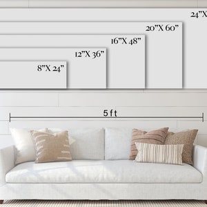 May include: A white sofa with cushions in a living room setting. The wall behind the sofa has five different sized rectangular frames with measurements in inches. The wall is white with horizontal wood panelling. The floor is a light brown wood. The sofa is 5 feet wide.