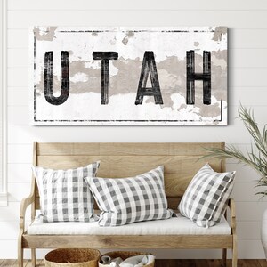 May include: A distressed white sign with black lettering that reads "UTAH". The sign is hanging on a white wall above a wooden bench with two checkered pillows.