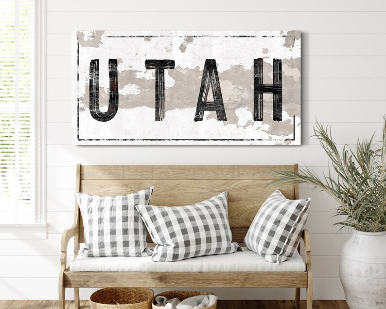 May include: A distressed white sign with black lettering that reads "UTAH". The sign is hanging on a white wall above a wooden bench with two checkered pillows.