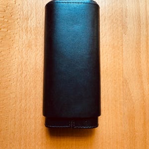 May include: A black leather cigar case. The rectangular case has a rounded top and a separate, smaller bottom section. The case is made of smooth, dark leather and is closed. The case is sitting on a light brown wooden surface.
