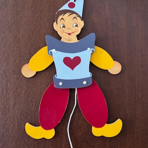May include: A wooden pull-string toy in the shape of a smiling boy. The boy has a blue hat with red dots, a blue and gray top with a red heart, yellow arms, and red legs with yellow shoes. A red string is attached to the top.