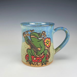 May include: A blue and brown ceramic mug with a green frog playing a banjo. The frog is sitting on a red mushroom with a yellow flower behind it.