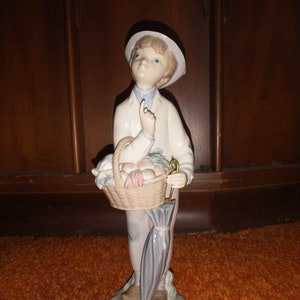 May include: A porcelain figurine of a boy wearing a white hat and holding a basket of fruit and an umbrella.