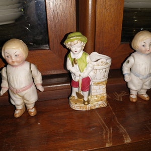May include: Three porcelain figurines of children. The figurine on the left is a boy wearing a white shirt, pink pants, and black shoes. The figurine in the middle is a boy wearing a green shirt, red pants, and black shoes. The figurine on the right is a boy wearing a white shirt, blue pants, and black shoes.