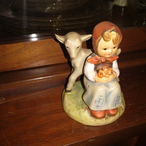 May include: A porcelain figurine of a girl in a red and white dress holding a basket of flowers. A white lamb stands beside her. The figurine is on a green base.