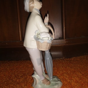 May include: A porcelain figurine of a boy wearing a white hat and holding a basket of flowers. He is standing on a green base with a blue umbrella.