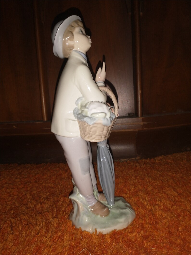 May include: A porcelain figurine of a boy wearing a white hat and holding a basket of flowers. He is standing on a green base with a blue umbrella.