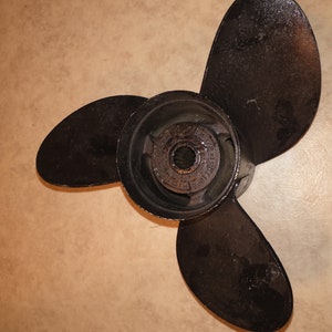 Könnte beinhalten: Ein dreiblättriger Propeller mit schwarzer Metalloberfläche. Der Propeller hat eine zentrale Nabe mit einer kreisförmigen Öffnung und einer Reihe kleiner Löcher am Rand.