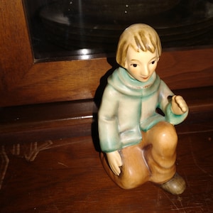 Może przedstawiać: Ceramiczna figurka przedstawiająca młodą osobę siedzącą z dłońmi złożonymi na kolanach. Figurka ma na sobie niebieską kurtkę i brązowe spodnie.