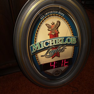 May include: A round, black framed clock with a gold trim. The clock face is blue with a white label that says "Michelob Light" and an eagle logo. The clock face also says "All Imported Hops". The clock has a red digital display showing the time 4:12.