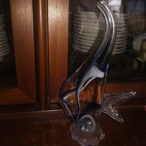 May include: A glass sculpture of a blue and white fish with a clear base. The fish is abstract and has a long, curved tail.