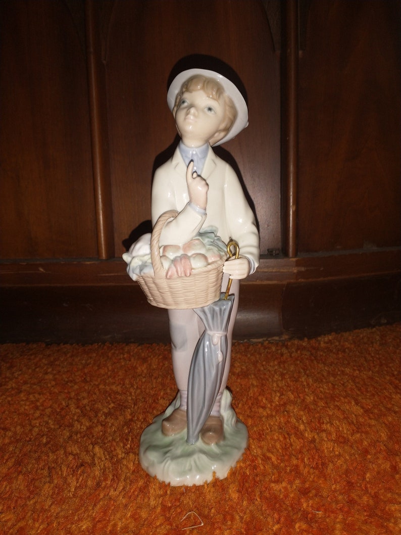 May include: A porcelain figurine of a boy wearing a white hat and holding a basket of fruit and an umbrella.