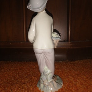 May include: A porcelain figurine of a girl wearing a white dress and a light blue hat, holding a basket of flowers. The figurine is standing on a green base.
