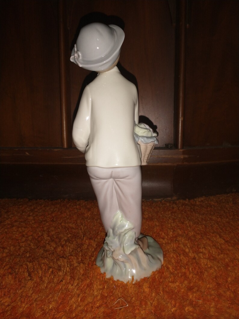 May include: A porcelain figurine of a girl wearing a white dress and a light blue hat, holding a basket of flowers. The figurine is standing on a green base.