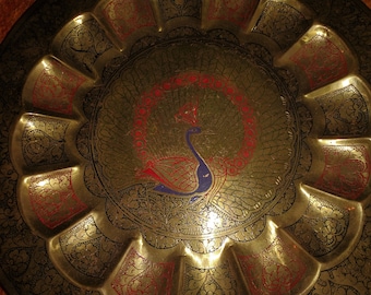 May include: A brass serving tray with a peacock design. The peacock is blue, red, and gold. The tray has a scalloped edge and intricate floral designs.