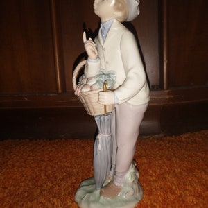 May include: A porcelain figurine of a young boy wearing a white jacket and a hat, holding a basket of fruit and an umbrella.