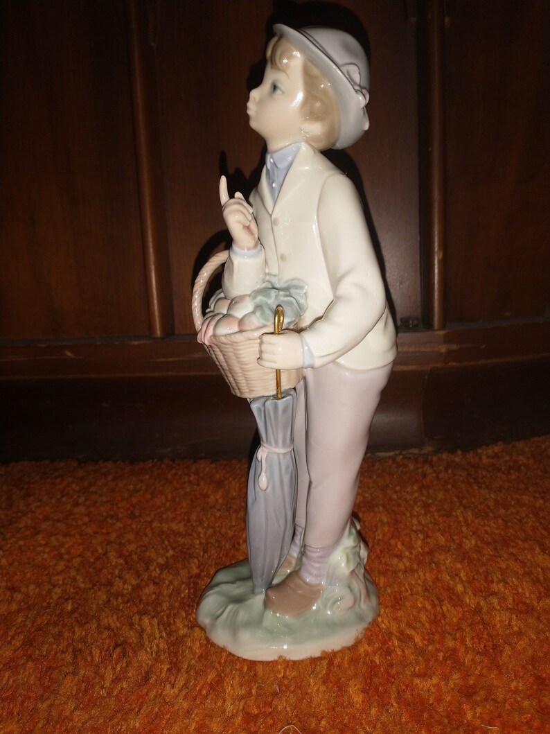 May include: A porcelain figurine of a young boy wearing a white jacket and a hat, holding a basket of fruit and an umbrella.