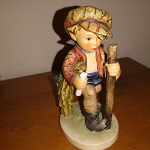 May include: A figurine of a young boy wearing a brown and green cap, a red and brown jacket, blue shorts, and black boots. He is holding a stick in his right hand and a white object in his left hand. He is standing on a green base.