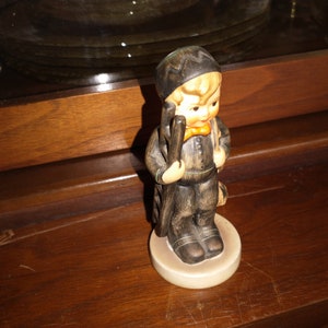 Może przedstawiać: Mała ceramiczna figurka chłopca przebranego za kominiarza. Ma na sobie czarny kapelusz i czarno-brązowy uniform. Trzyma szczotkę i torbę. Figurka stoi na okrągłej podstawie.