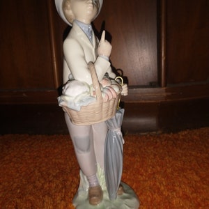 May include: A porcelain figurine of a boy wearing a black hat and a white jacket, holding a basket of fruit and an umbrella. The figurine is standing on a green base.