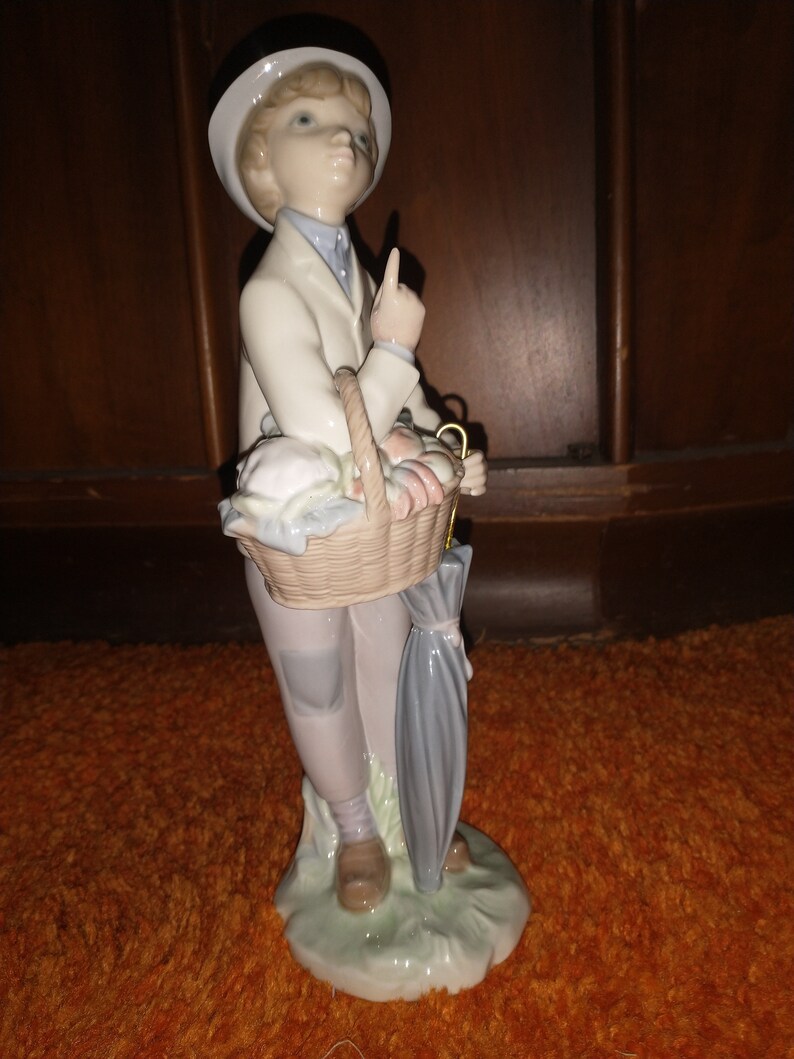 May include: A porcelain figurine of a boy wearing a black hat and a white jacket, holding a basket of fruit and an umbrella. The figurine is standing on a green base.