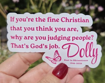 May include: A white die-cut sticker with pink text that reads: If you're the fine Christian that you think you are, why are you judging people? That's God's job. Dolly, Rest in Rhinestones, 1946-2026. A small pink butterfly graphic is included.