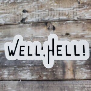 May include: White sticker with black text that reads "WELL, HELL!" The sticker is on a wooden surface.