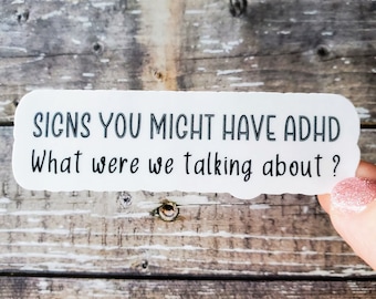 May include: A white sticker with black text that reads "SIGNS YOU MIGHT HAVE ADHD" and "What were we talking about?". The sticker is held by a person with a pink glitter nail polish. The background is a weathered, wooden surface.