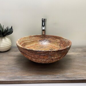 May include: A round, natural stone vessel sink with a polished chrome faucet. The sink has a layered, earthy color pattern of browns, tans, and creams. A small, white vase with a faux succulent sits to the left.