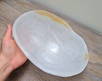 May include: A white and translucent onyx bowl with a kidney bean shape. The bowl has a smooth surface with natural veining and a golden-yellow rim. The bowl is held in a person's hand.