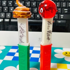 May include: Two PEZ dispensers. One features a yellow and brown snake head, the other a red head with a face. Both have white stems with text and are on green and red bases.