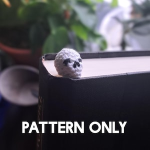 May include: A small, crocheted skull in light gray yarn with black facial features. The skull is perched on the edge of a book. The text "PATTERN ONLY" is displayed in white block letters at the bottom of the image.