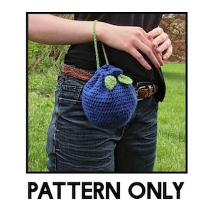 May include: A crocheted blueberry-shaped bag with green leaves. The bag is attached to a brown leather belt and is being worn.