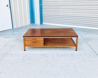 May include: A rectangular wooden coffee table with a warm brown finish. It features a lower shelf and two drawers on the left side. The table has a black trim around the top and stands on four legs with black feet. The table measures approximately 48 inches long.