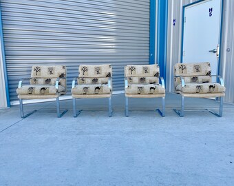 May include: Four chrome and fabric chairs with a patterned design of trees and camels. The chairs are arranged in a row on a concrete surface.
