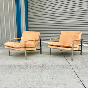 May include: Two mid-century modern armchairs. The chairs have a light orange fabric upholstery and chrome frames. The armrests are made of wood. The chairs are placed on a concrete surface in front of a gray garage door.