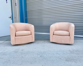 May include: Two blush-colored swivel chairs with rounded backs and cushioned seats. The chairs are made of a textured fabric and sit on a wooden base. The chairs are placed on a concrete surface.