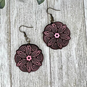 Puede incluir: Un par de pendientes florales rosas y marrones. Los pendientes presentan un diseño circular con una flor rosa en el centro y detalles rosas. Cuelgan de ganchos para las orejas de color bronce.