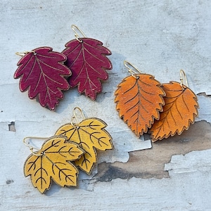 May include: Three pairs of leaf-shaped earrings with gold-toned metal hooks. One pair is deep burgundy, one pair is bright orange, and one pair is mustard yellow. Each earring features detailed etched vein patterns on the surface of the leaf shape.