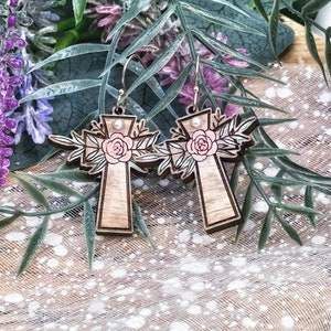 May include: A pair of wooden cross earrings with a pink floral design and a pearl accent. The earrings are made of light brown wood and have a delicate floral pattern.
