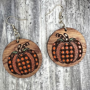 Puede incluir: Un par de pendientes redondos de madera con un diseño de calabaza. Las calabazas son de color naranja con un patrón de cuadros negros y blancos y tallos verdes.