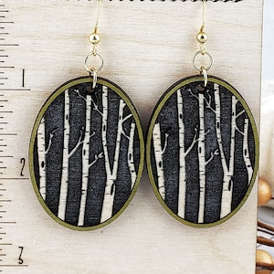 May include: A pair of round wooden earrings with a black and white birch tree design. The earrings have a gold-colored hook and a green border.