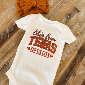 May include: A white short-sleeved baby bodysuit featuring the orange glitter text "She's from TEXAS I CAN TELL" with a longhorn graphic and two small hearts. A matching burnt orange cable-knit fabric headband with a large bow is included.